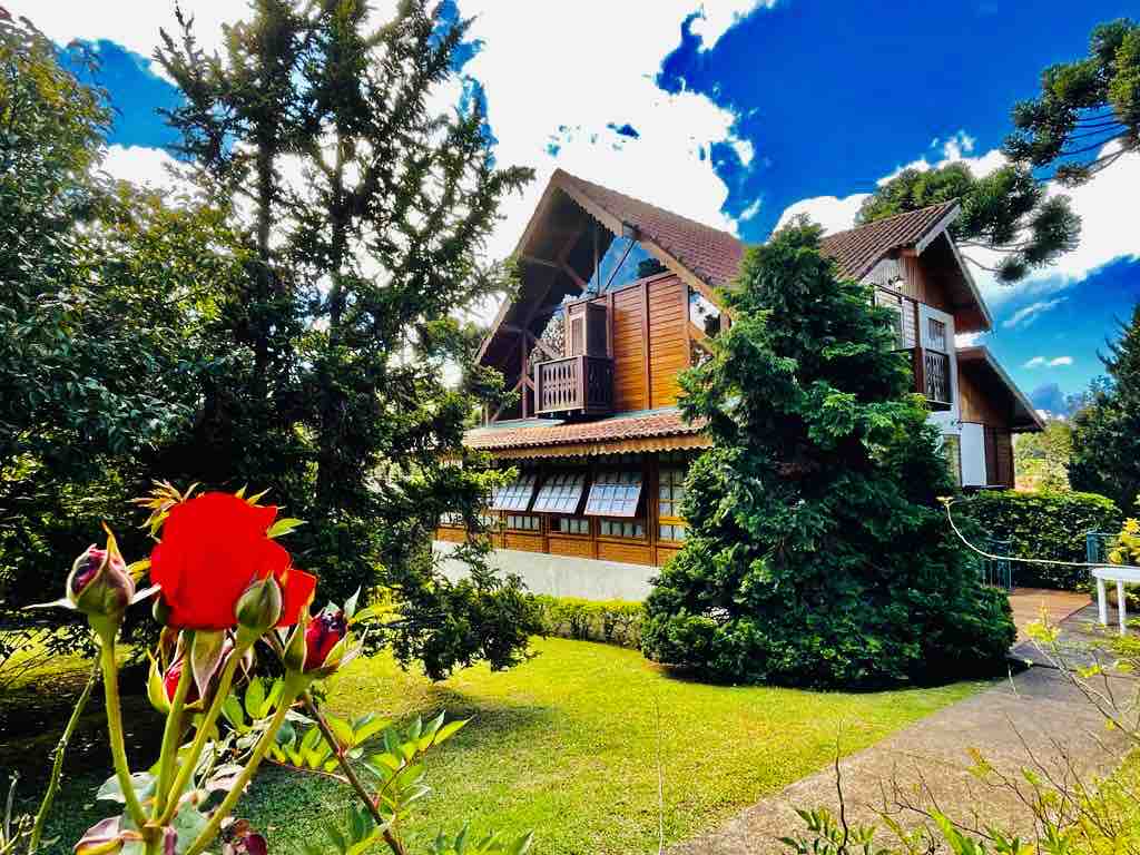 A charming two-story chalet is surrounded by lush greenery and vibrant flower beds. The wooden structure features a gabled roof and large windows, allowing light to fill the interior. A bright red rose in the foreground adds a touch of color to the serene outdoor setting.