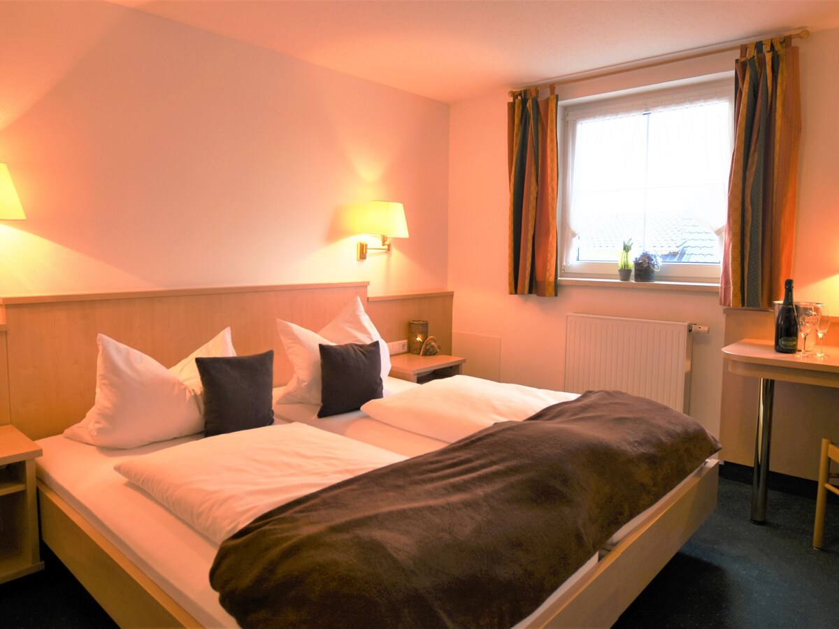 A cozy room features a spacious bed with pillows and soft linens, complemented by warm wall lighting. A window allows natural light to enter, framed by curtains. A small table with two chairs is positioned near the wall, enhancing the room's functional layout.