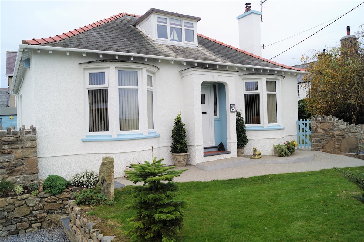 The exterior of a charming white house features blue accents and a well-maintained garden. A neatly paved pathway leads to the front door, surrounded by low stone walls and plants. Large windows allow for ample natural light, contributing to an open and inviting appearance.