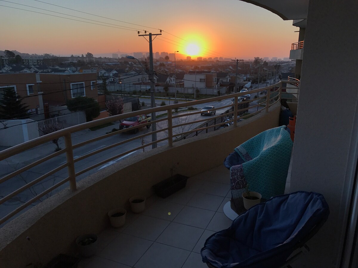 A private balcony offers a view of a sunset over the neighborhood, with soft orange and pink hues illuminating the sky. Two chairs are positioned on the tiled floor, complemented by potted plants. The railing provides a clear sightline to the street and surrounding area.
