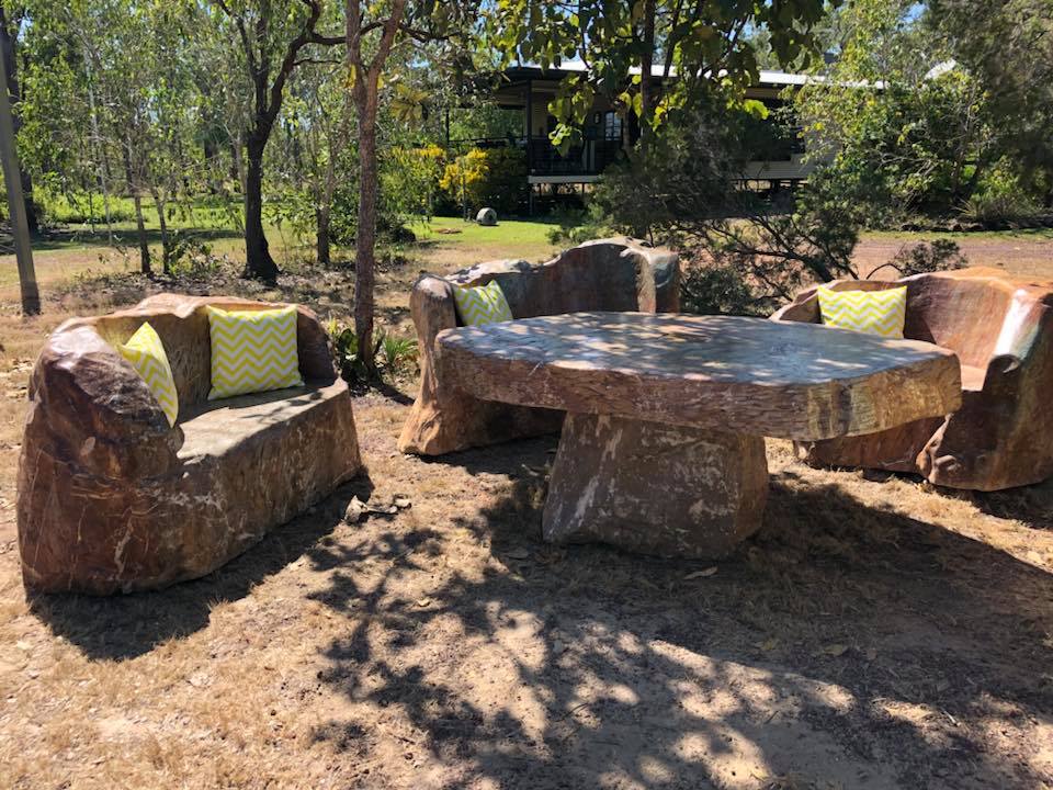 A set of rustic outdoor furniture features a round table and two seating areas crafted from natural materials. The pieces have a raw, earthy appearance, complemented by bright yellow and green chevron-patterned cushions. Surrounding greenery enhances the serene outdoor setting.