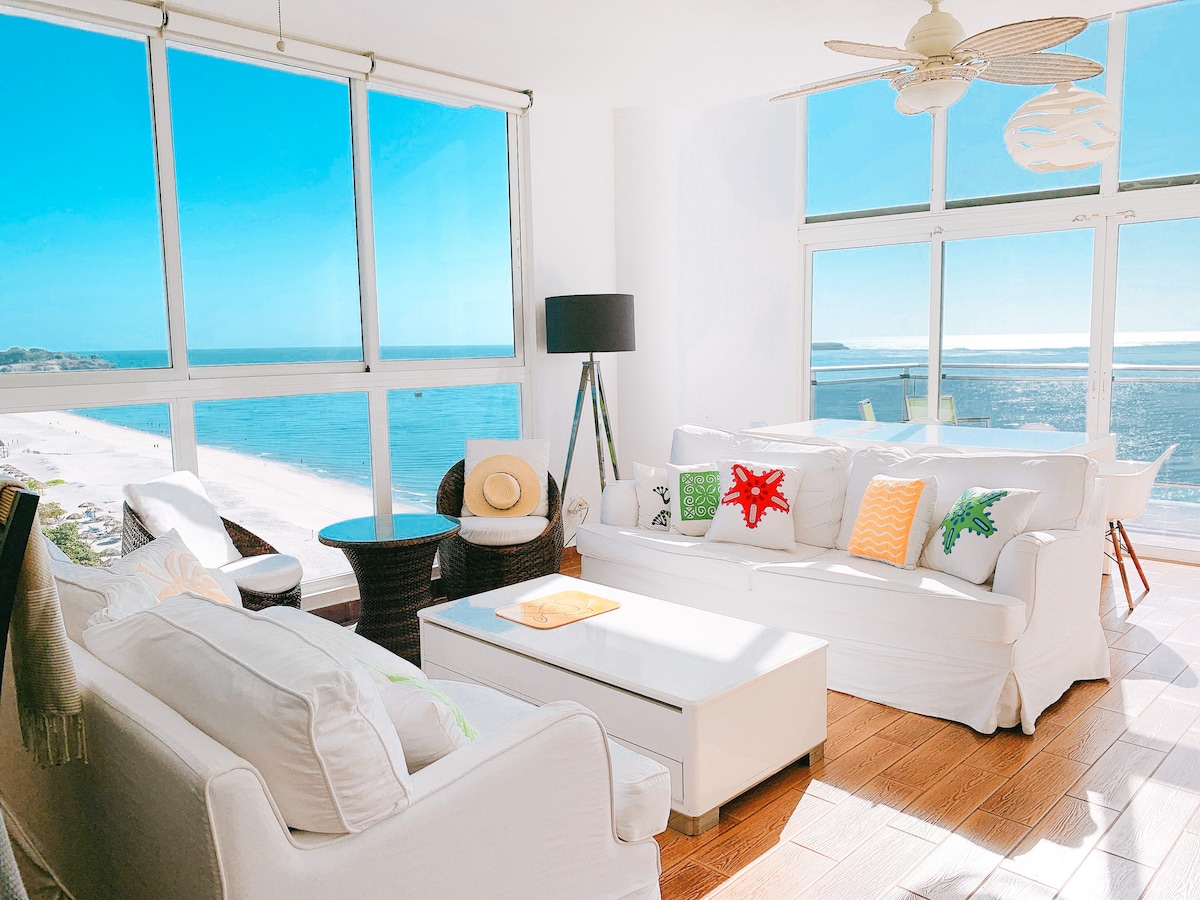 A bright living area features large glass windows that provide expansive views of the beach and ocean. White sofas are arranged around a minimalist coffee table, with decorative pillows adding subtle color. Natural light floods the space, enhancing its open and airy feel.