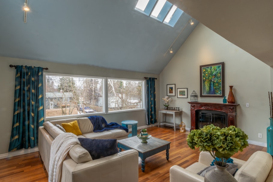 A bright, airy living room features large windows that allow natural light to illuminate the space. Two light-colored sofas face a decorative fireplace, while a coffee table sits in the center. Colorful curtains and artwork provide a vibrant contrast to the warm wooden flooring.
