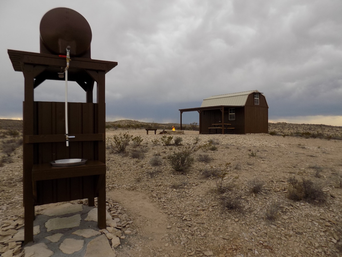 An outdoor shower is positioned next to the camping cabin, featuring a large water tank above a simple wooden structure. The cabin is seen in the distance, surrounded by rocky terrain and sparse vegetation. A fire pit and picnic area are slightly visible, indicating a tranquil outdoor setting.