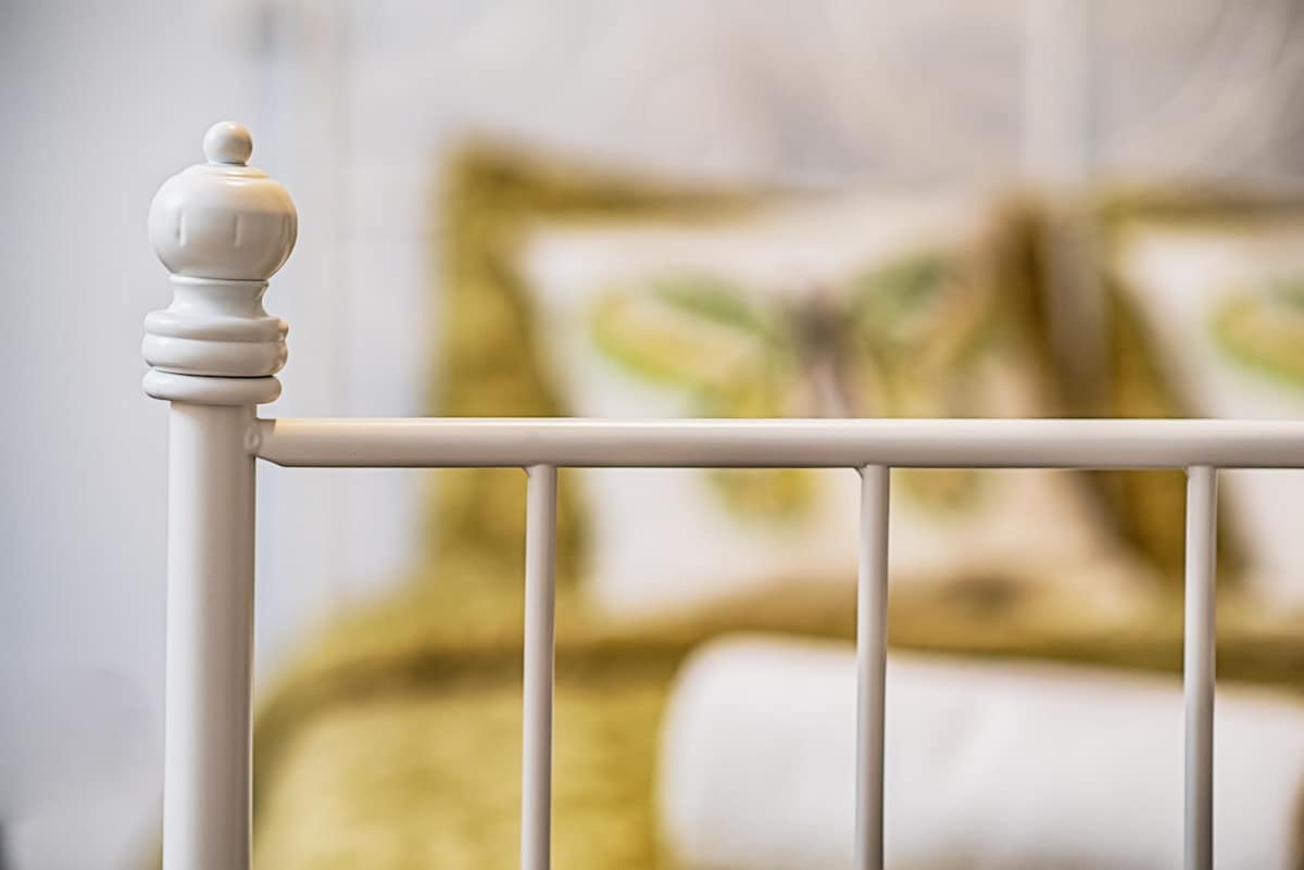 A close-up view of a white metal bed frame is presented, featuring a decorative finial. Soft bedding in muted tones is visible in the background, accented by patterned pillows. A rolled towel rests neatly beside the bed, adding to the inviting arrangement.