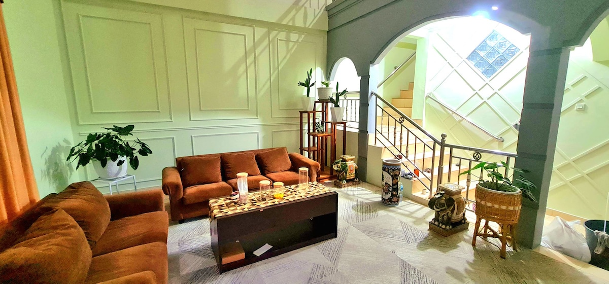 A spacious living area is furnished with two brown sofas and a low coffee table with decorative elements. Plants are strategically placed around the room, and a stylish staircase, featured in the background, leads to the upper level.