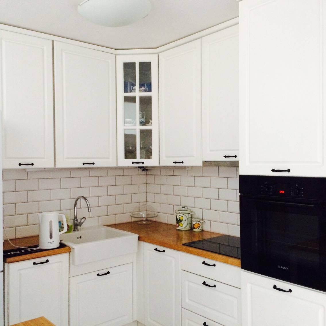 A modern kitchen features white cabinetry and a wooden countertop. A sink is located in one corner, with a kettle and appliances visible nearby. Glass-fronted cabinets display dishware, while a built-in oven and stovetop are situated along the wall.