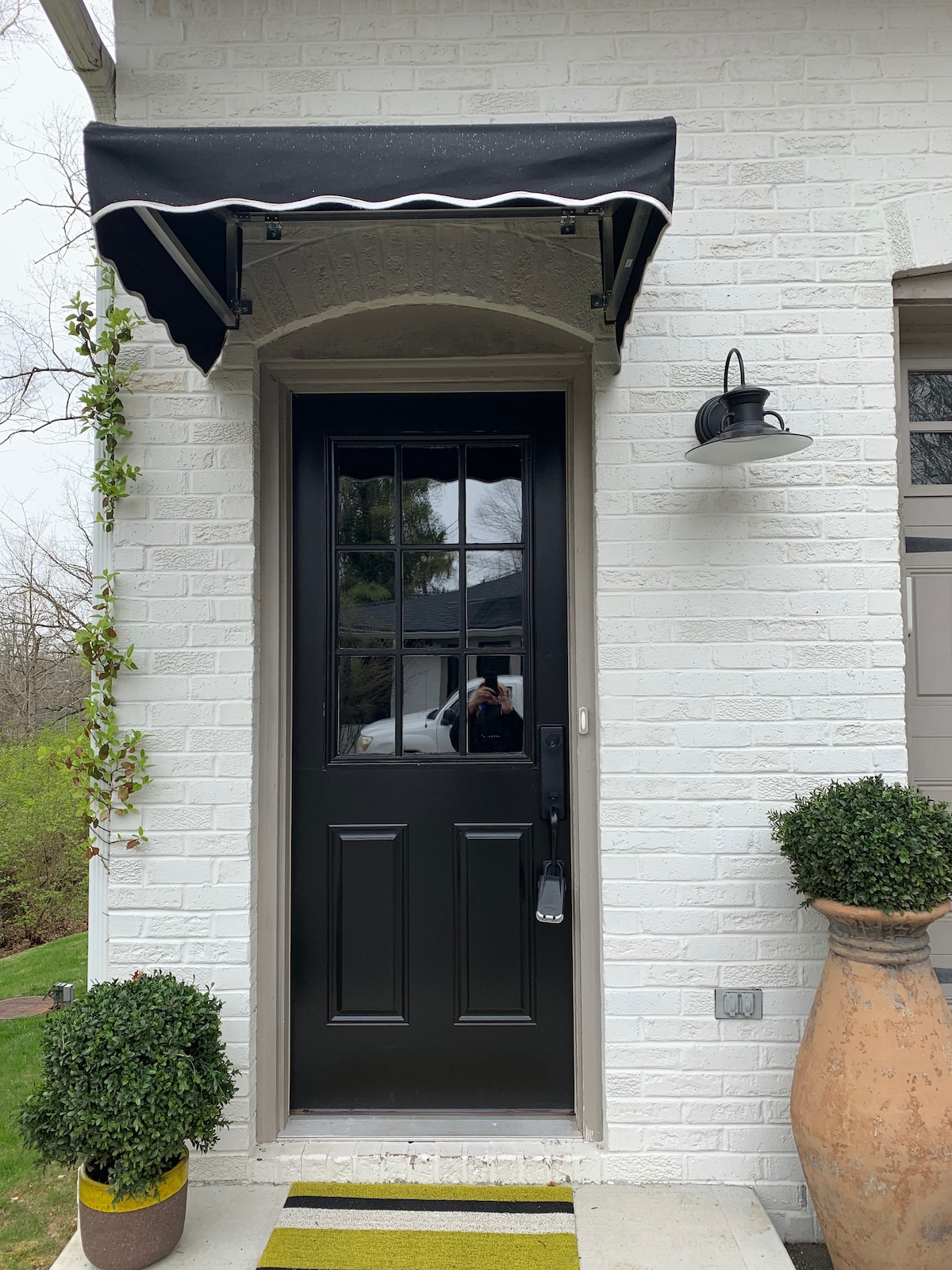 The entrance features a black door with glass panels, framed by a white-brick wall. Above, an awning provides shade, while a wall-mounted light fixture adds illumination. On either side, small potted plants create a welcoming appearance, complemented by a striped doormat.