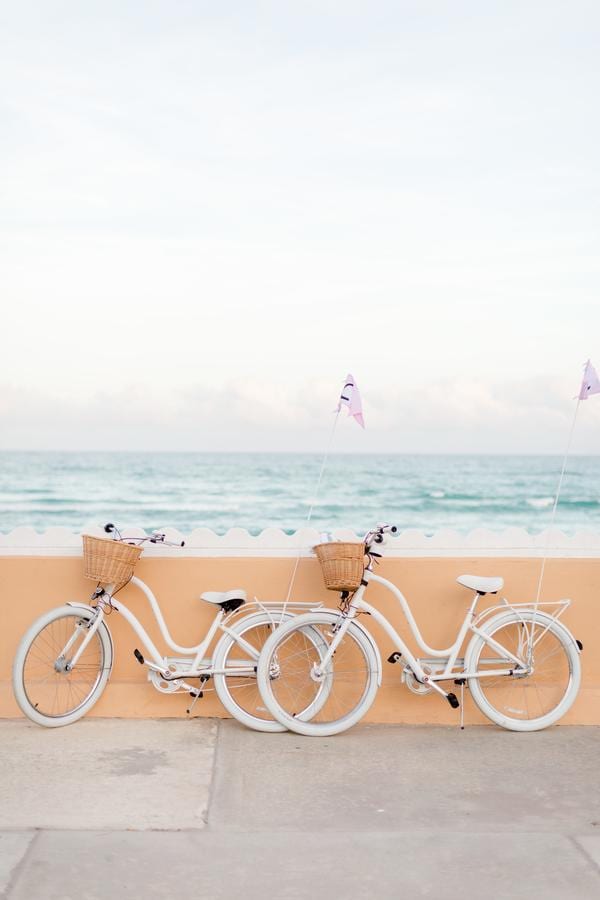Two white bicycles are positioned against a pastel-colored wall, each featuring a wicker basket attached to the front. The backdrop shows the tranquil ocean, with gentle waves lapping against the shore under a soft sky.