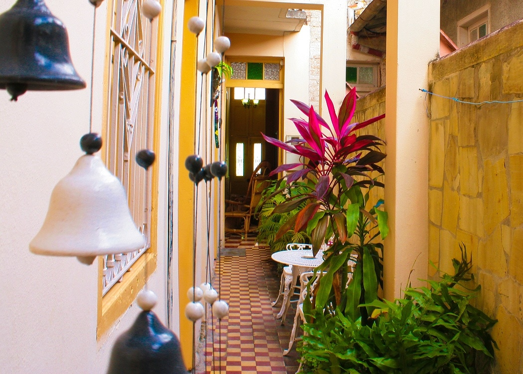A narrow corridor is adorned with decorative wind chimes and vibrant plants. A small white table with matching chairs is positioned along the side, inviting relaxation. The corridor features tiled flooring and walls painted in warm hues, creating an atmosphere of comfort and tranquility.