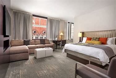 A spacious studio room features a king bed with decorative pillows and a large pullout sofa, accompanied by a modern desk and chair. Natural light enters through large windows, while neutral-toned curtains frame the space. A flat-screen television is mounted on the wall.