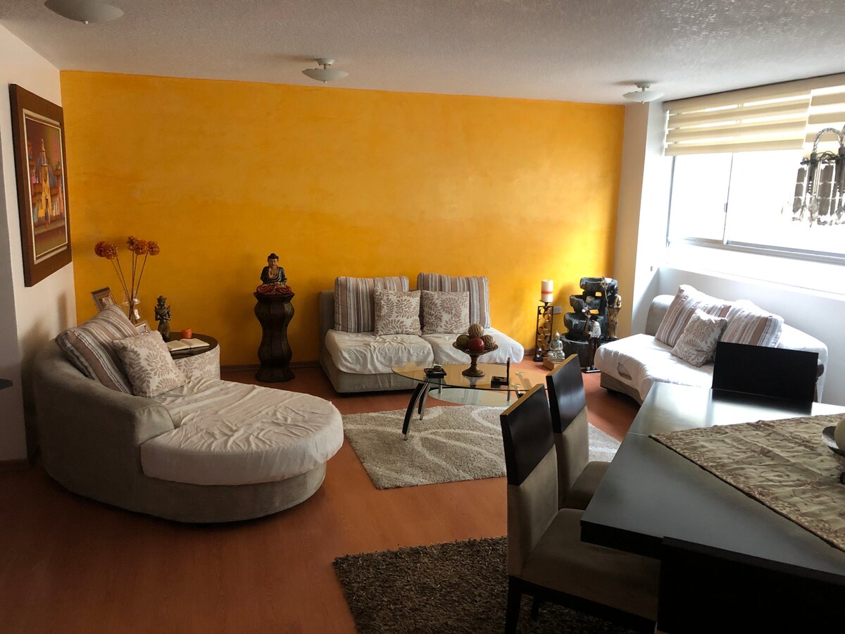 A welcoming living area features light-colored upholstered seating arranged around a glass coffee table. A vibrant yellow wall adds warmth to the space. Soft rugs are positioned underfoot, while large windows allow natural light to fill the room, complemented by modern light fixtures above.