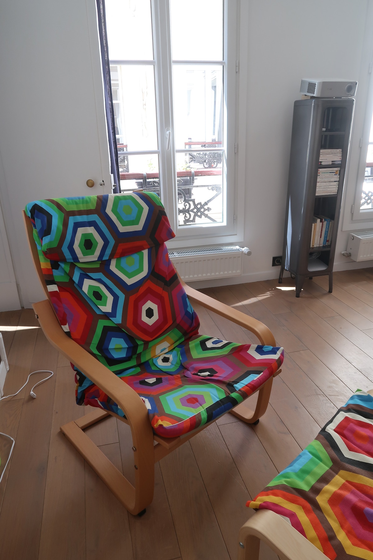 A colorful rocking chair with a hexagonal patterned fabric is positioned on warm wooden flooring. Large windows allow natural light to fill the space, highlighting the vibrant hues of the chair and creating a lively atmosphere.