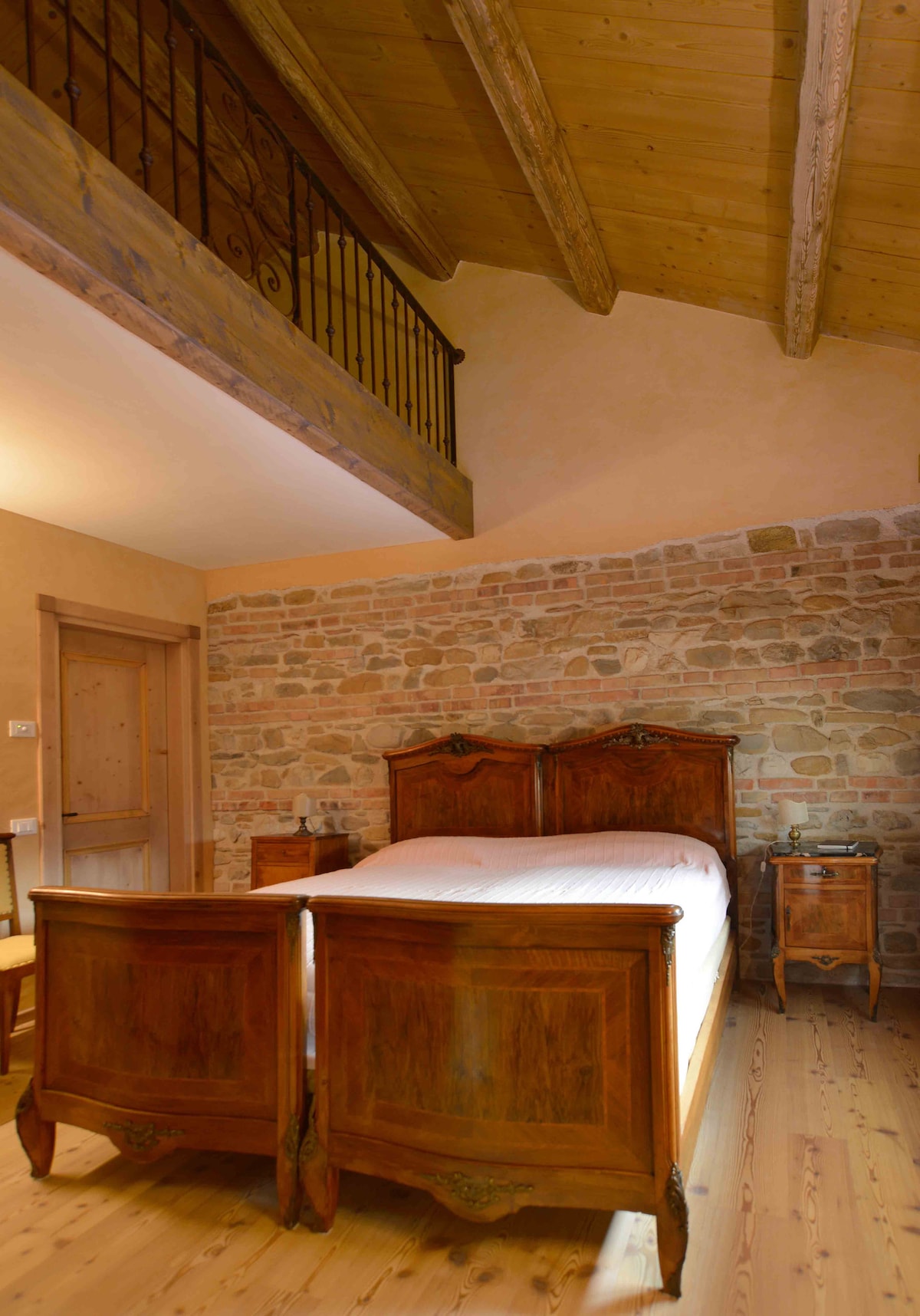 A spacious bedroom features a double bed framed by two ornate wooden headboards, set against a backdrop of warm stone walls. High wooden beams enhance the vertical space, while soft, ambient light casts a gentle glow throughout the room.