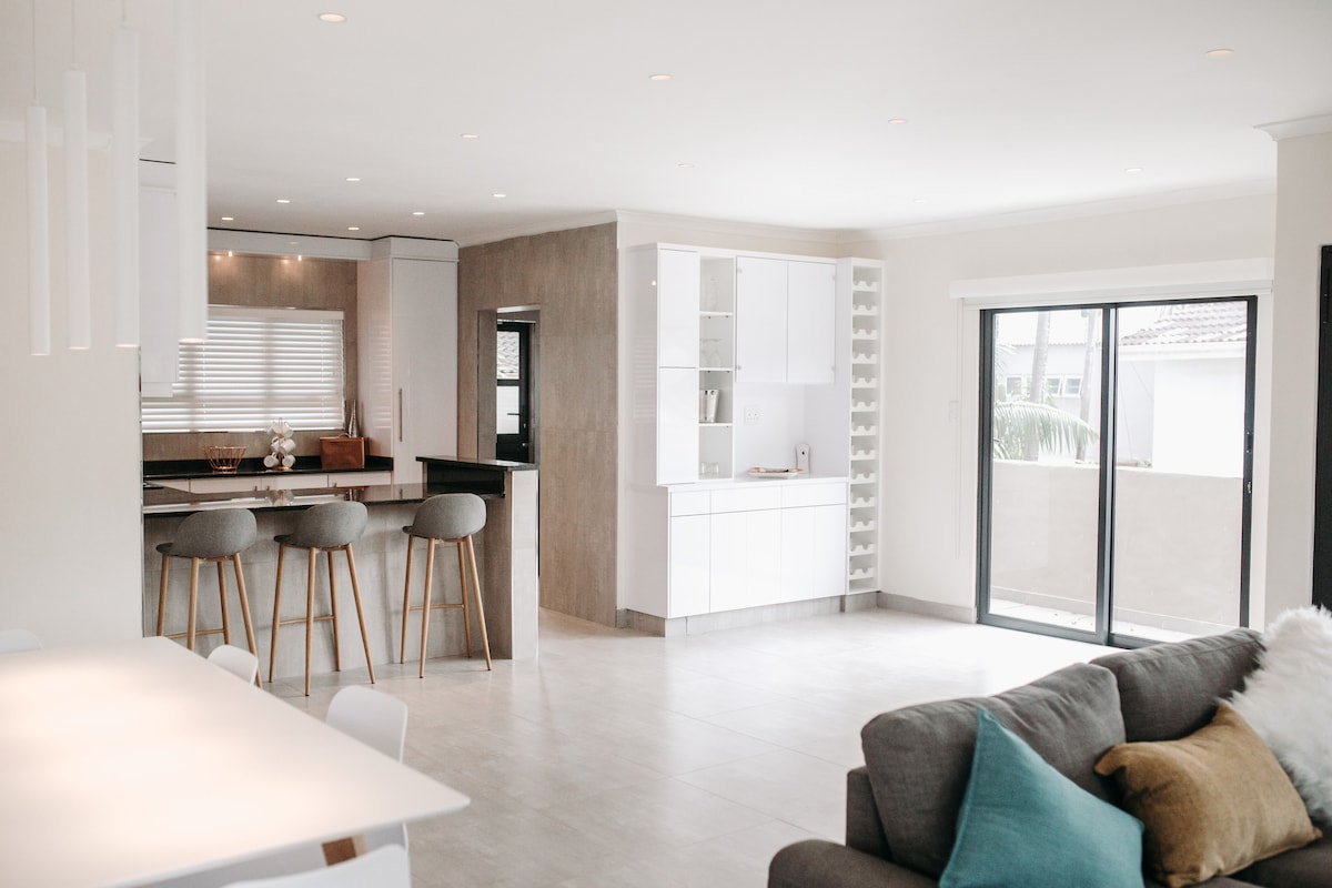 A spacious and modern living area features a light and neutral color palette, complemented by contemporary furnishings. A dining table is set for six, while bar stools line the kitchen counter. Wide sliding glass doors open to an outdoor space, enhancing natural light.