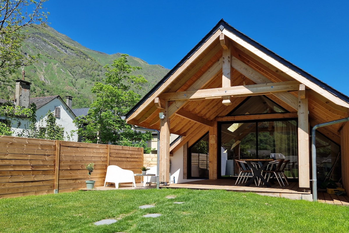 A modern chalet features a wooden structure with a sloped roof and large glass walls. A spacious terrace overlooks a landscaped garden with green grass and stepping stones. Nearby, a white chair is positioned beside a potted plant, offering a relaxed outdoor setting against a mountain backdrop.