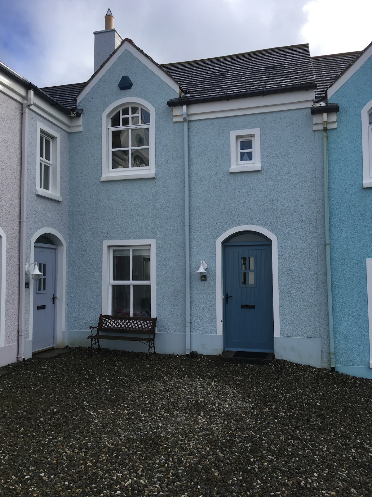 The exterior of the townhouse features a light blue façade, highlighted by white trim around the windows and doors. A small bench sits outside, positioned on a gravel area. The entrance door is painted in a complementary shade of blue, creating a cohesive look.