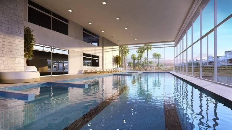 An indoor swimming pool area features multiple lanes and lounge seating surrounded by large glass windows. Natural light floods the space, highlighting a calm water surface that reflects the modern architecture and indoor greenery. Palm trees are visible outside, adding a serene touch.