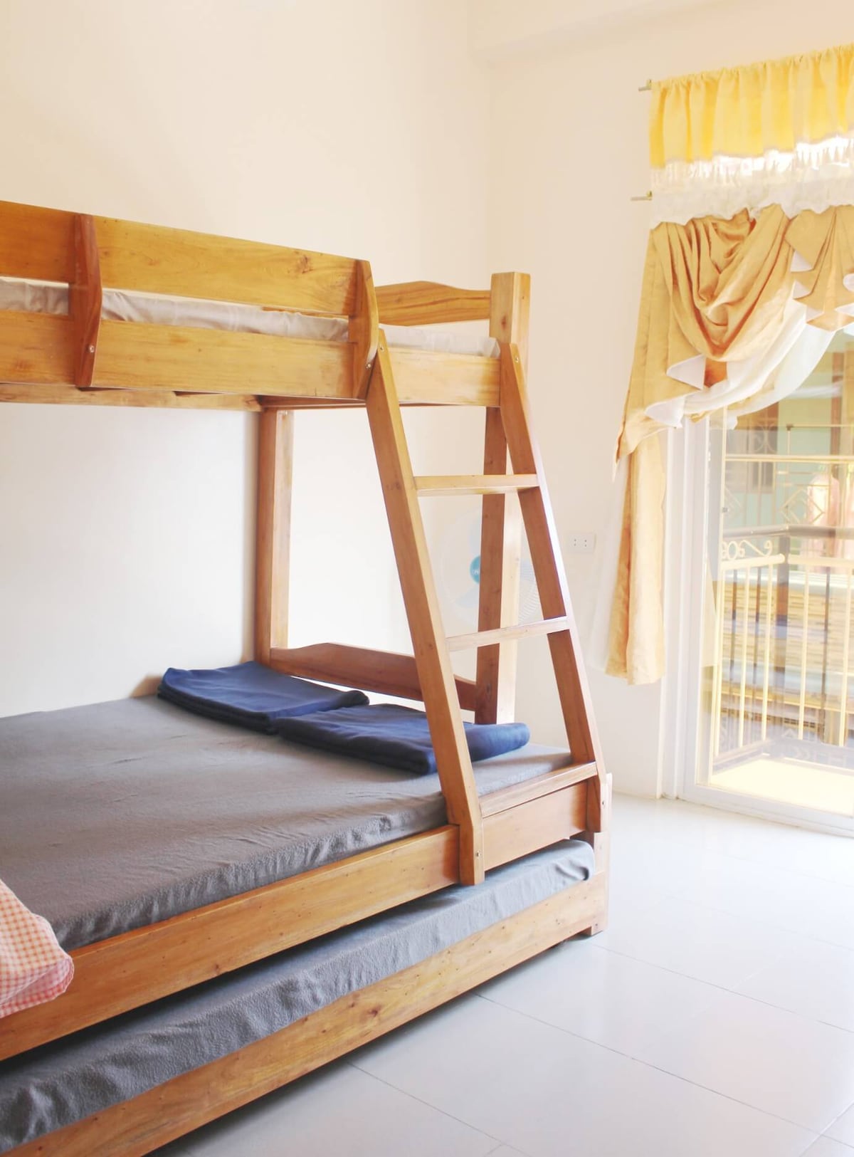 A wooden bunk bed is featured in this room, with a lower mattress neatly made and a thin blanket placed on it. Light is coming through a window, accompanied by yellow drapes that add a soft touch to the bright space.