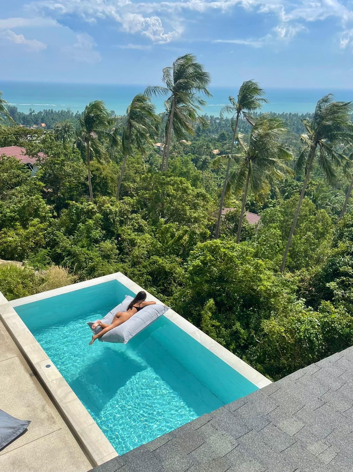 An infinity pool stretches towards a scenic view of palm trees and the ocean. The pool, featuring clear blue water, includes a cushioned lounge area. Sunlight reflects off the water, enhancing the serene environment surrounded by lush greenery.