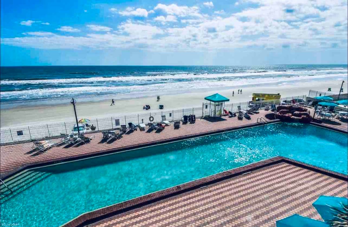 The image features a clear view of the oceanfront, showcasing the sandy beach and Atlantic Ocean. A sparkling outdoor pool is visible in the foreground, surrounded by lounge chairs. Bright blue skies punctuated by fluffy clouds create a cheerful atmosphere.