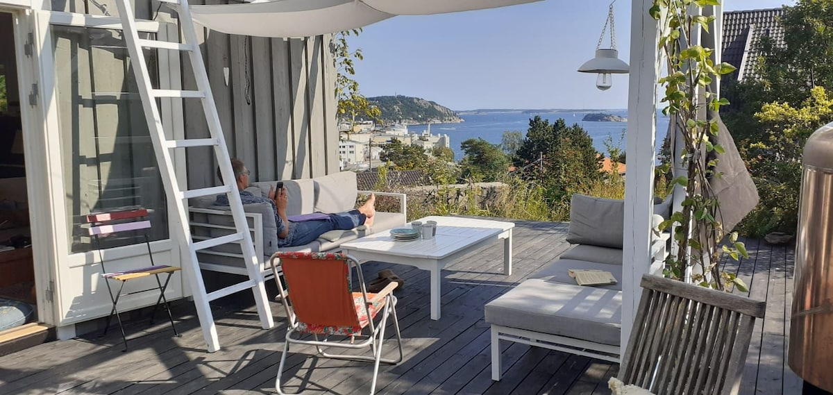 An outdoor terrace is presented with a partial view of the fjord, featuring seating areas and a coffee table. The space is adorned with greenery and a hanging light fixture above. A relaxing atmosphere is created by the comfortable lounge chairs and a separate table for outdoor dining.