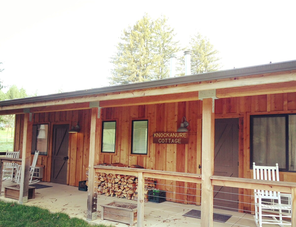 The exterior of Knockanure B features a warm wooden facade with a porch area. Rocking chairs are positioned beside a stack of firewood. Large windows allow natural light to illuminate the entrance, which includes two doors leading into the cozy cabin.
