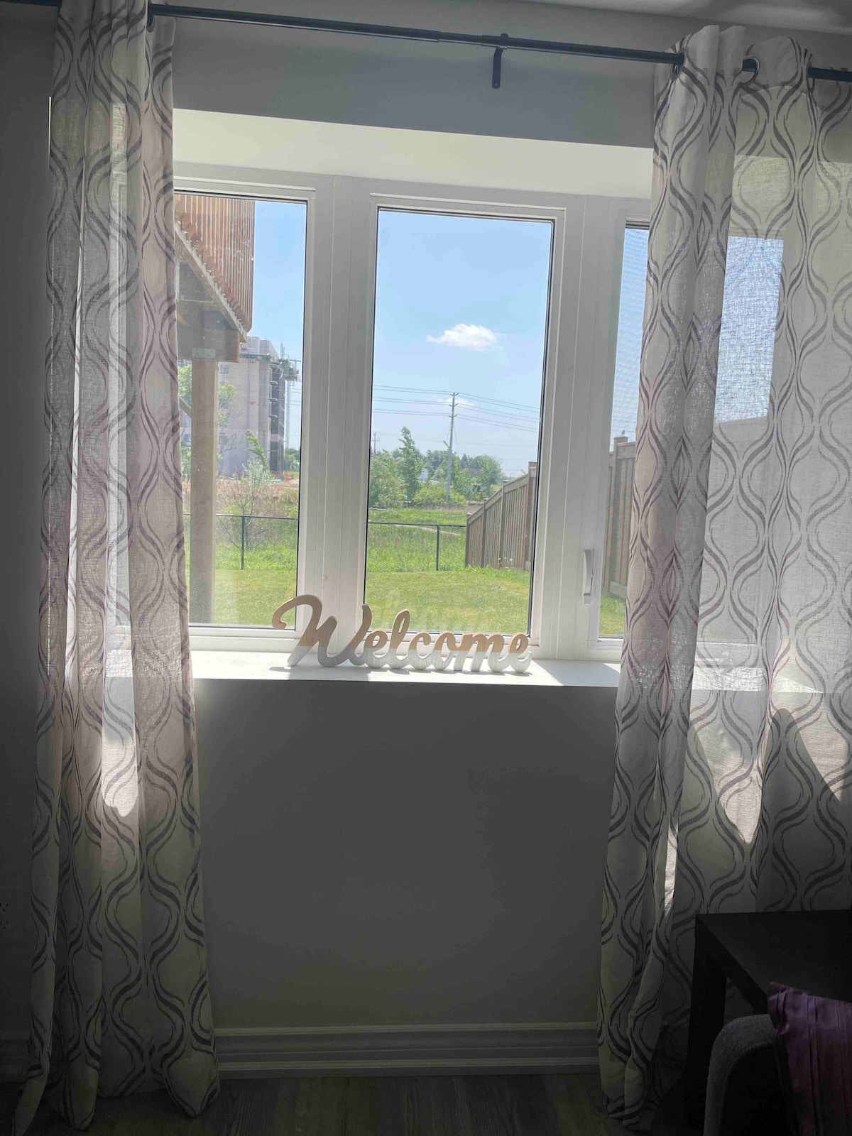 A bright window is framed by flowing sheer curtains, allowing natural light to fill the space. A decorative 'Welcome' sign rests on the windowsill, while an outdoor view of greenery and clear skies is visible beyond the glass.