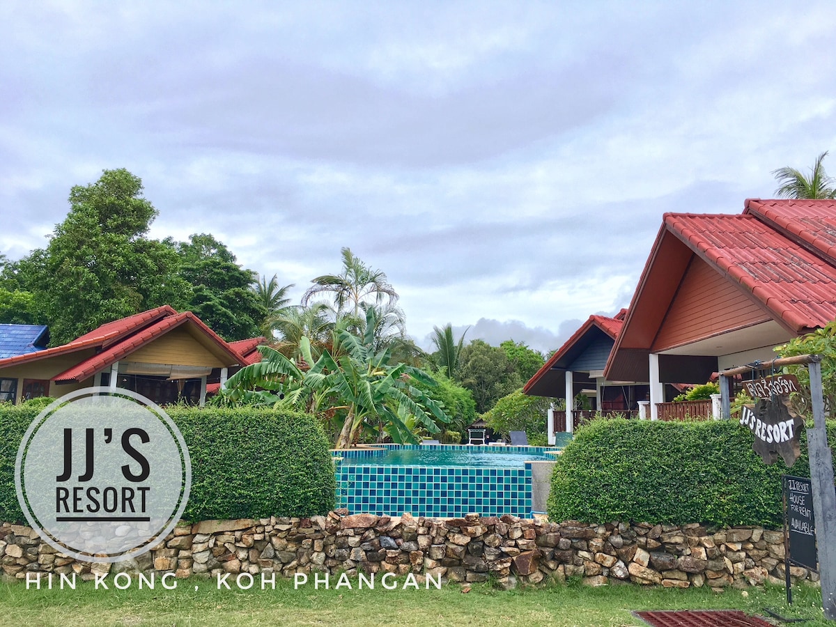 The image depicts a serene view of JJ's Resort, featuring bungalow-style accommodations surrounded by lush greenery. A turquoise pool occupies the center, framed by tropical plants and inviting outdoor spaces under a cloudy sky.