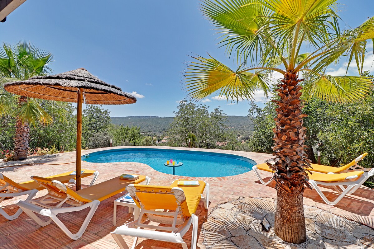 A serene outdoor area features a sparkling blue pool surrounded by sun loungers. Tropical palm trees are visible, offering shade with a thatched umbrella. The view extends into the lush landscape, providing a peaceful backdrop for relaxation.