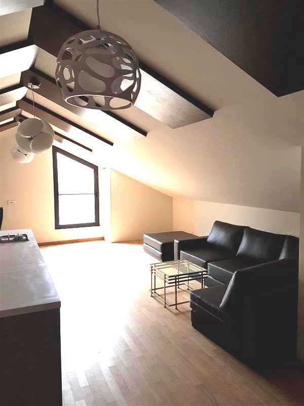 A bright living area is highlighted by angled ceilings and large windows, offering natural light. Two black leather chairs and a contemporary glass table provide seating. Simple light fixtures hang from the ceiling, adding a modern touch to the space.