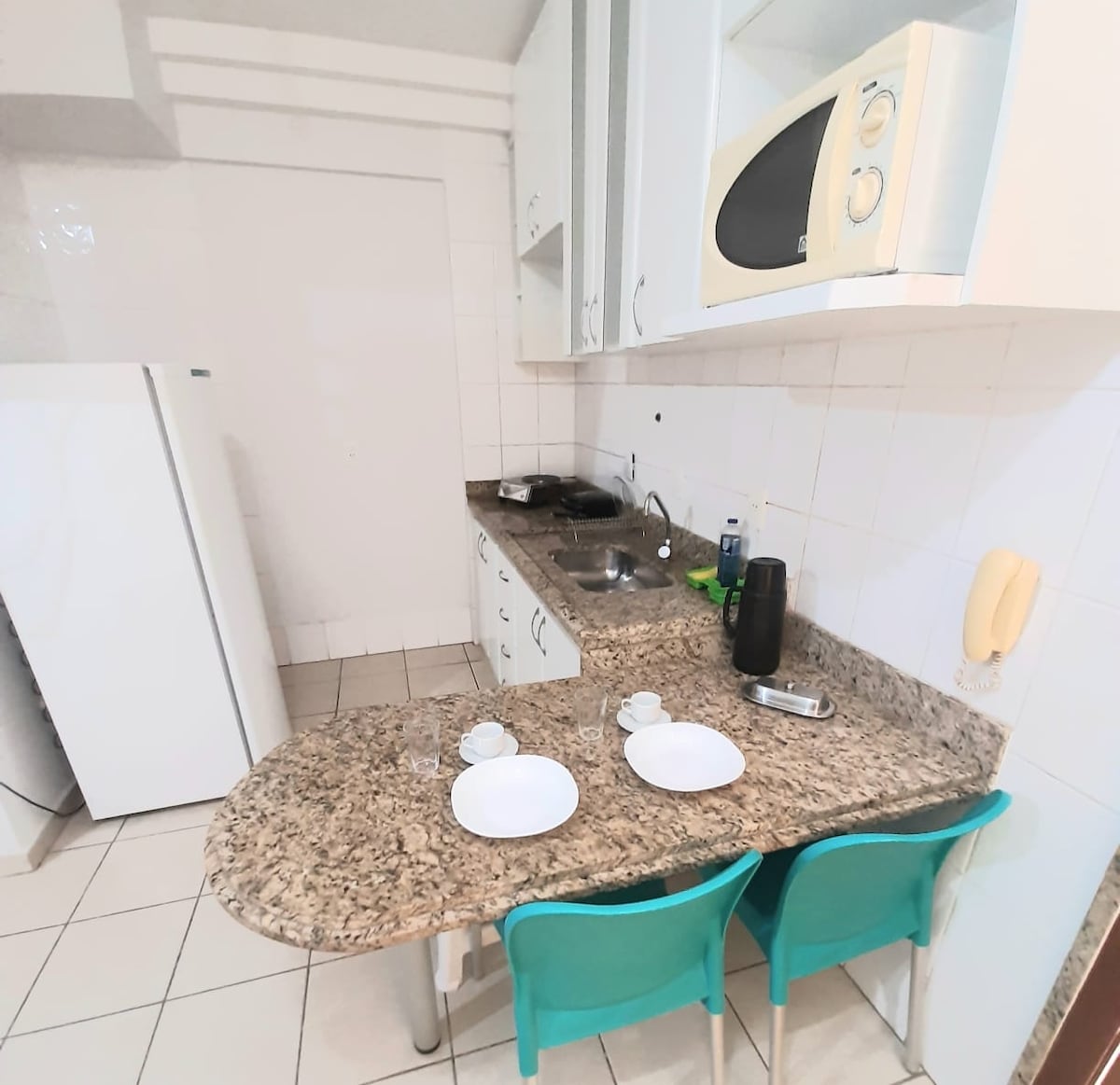 A compact kitchen space features a granite countertop with two plates and cups set for dining. Cabinets line the walls, and essential appliances, including a microwave and sink, are arranged for convenience. The tiled floor adds a clean, functional touch.