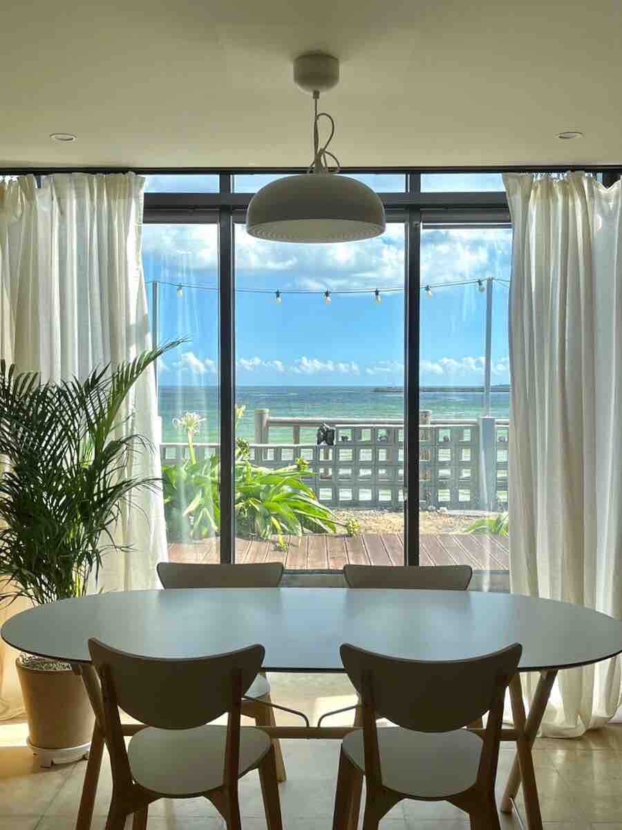 A bright dining area is showcased with a round table and four chairs. Large glass doors open to a balcony with a view of the ocean. Light curtains frame the windows, and a potted plant adds greenery to the space.