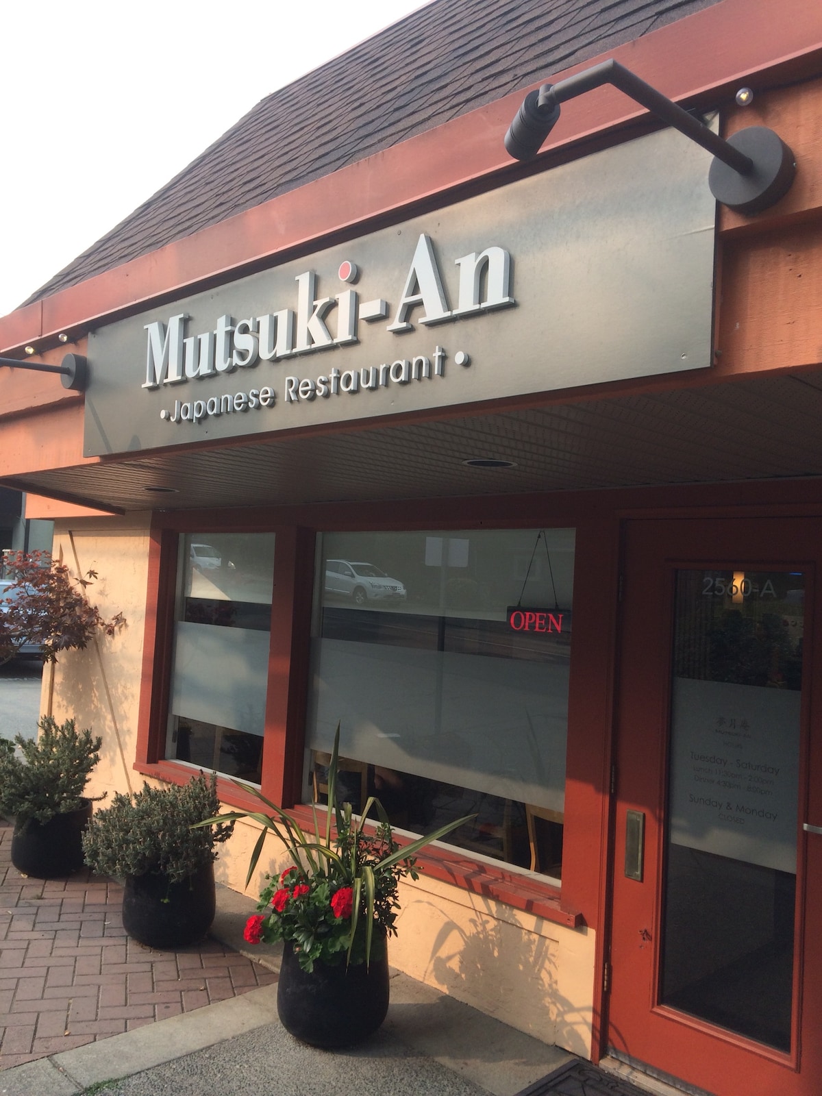 The exterior of a Japanese restaurant is displayed, featuring a prominent sign that reads 'Mutsuki-An.' Large glass windows provide a view inside, while potted plants with vibrant flowers decorate the entrance. An 'OPEN' sign is illuminated, welcoming guests.