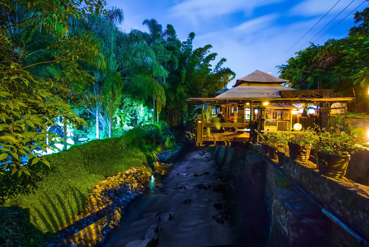 A charming cabin is nestled by a gentle stream, surrounded by lush greenery. Soft lighting illuminates the outdoor seating area and pathways, while tropical plants create a serene backdrop. The sound of flowing water enhances the tranquil setting, ideal for relaxation and rejuvenation.