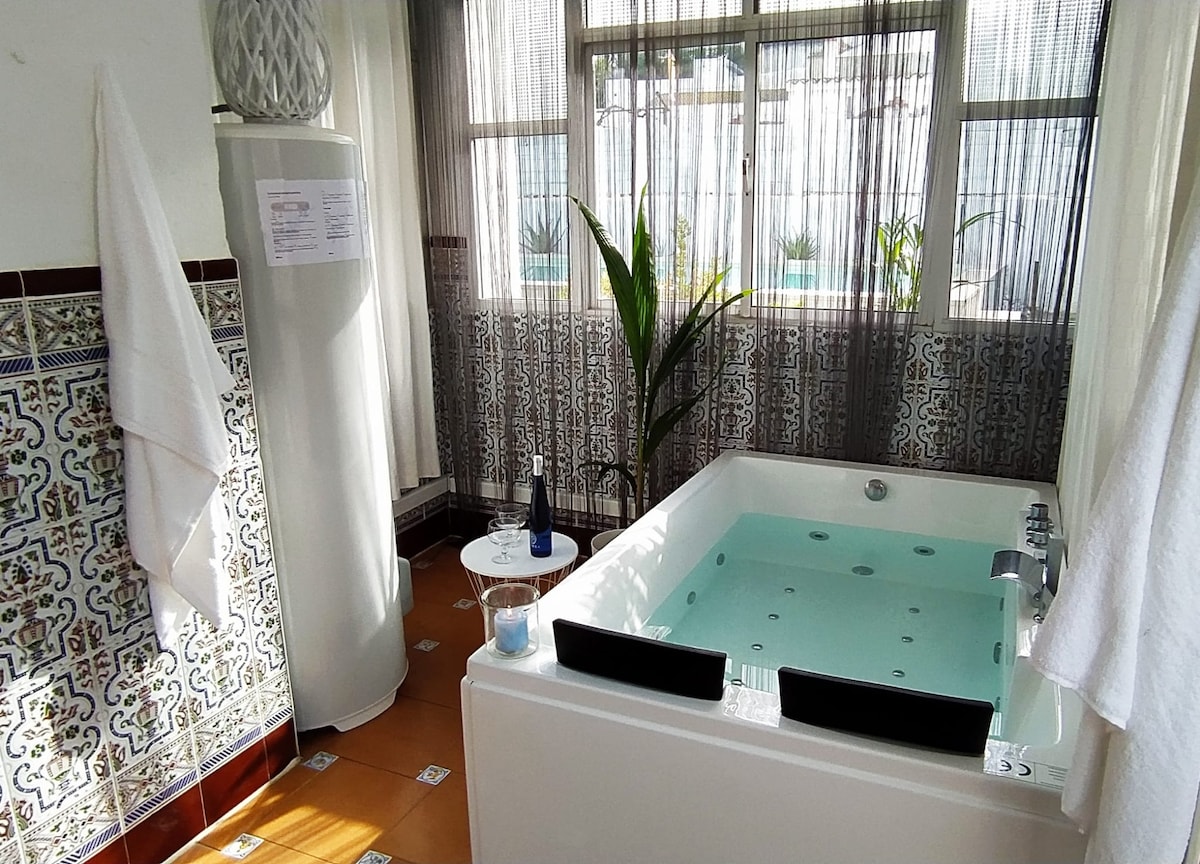 A bright and airy space features a private jacuzzi, complemented by light-colored tiles and glass windows that allow natural light. White towels are neatly displayed on a nearby rack, and a plant adds a touch of greenery to the setting.