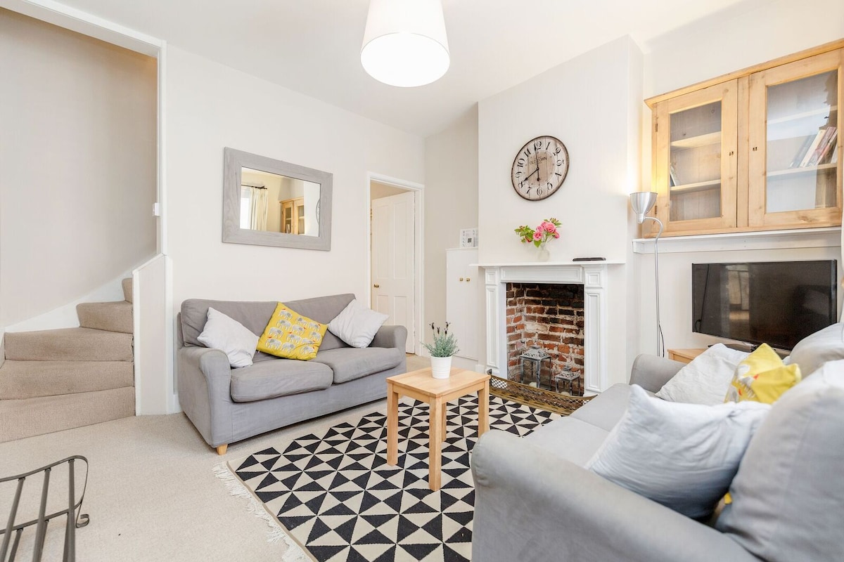 A sitting room features two grey sofas arranged around a light wooden coffee table on a patterned rug. A decorative fireplace with candles and a wall clock adds character. A staircase is visible, and natural light fills the space through large windows.