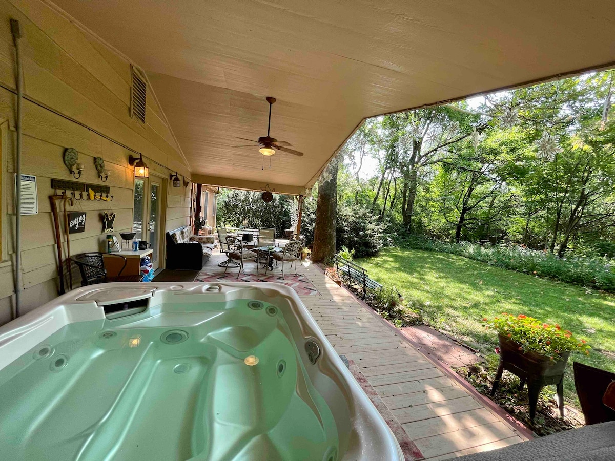 A spacious covered deck features a hot tub, inviting relaxation while surrounded by greenery. Comfortable seating and a dining area are positioned nearby, with views of the lush backyard offering a natural retreat. The area is well-lit and offers easy access to the outdoor garden.