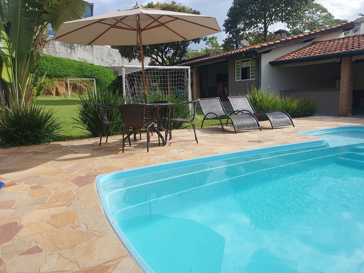 A serene pool area features clear blue water surrounded by a stone patio. Sun loungers are positioned nearby under an umbrella. A grassy field with a soccer goal is visible in the background, complementing the relaxed outdoor setting.