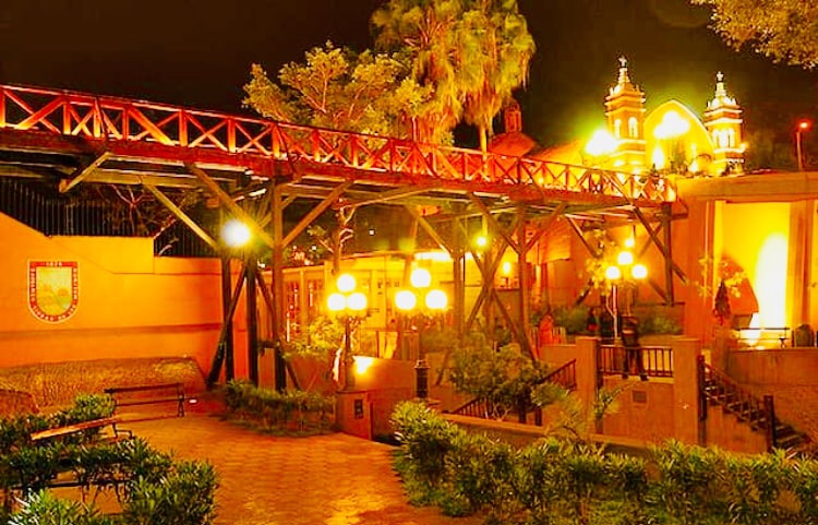 A walkway constructed of wooden beams leads through a well-lit garden area, surrounded by lush greenery. Antique-style lanterns hang, casting a soft glow. In the background, illuminated architecture adds a warm ambiance to the outdoor space.