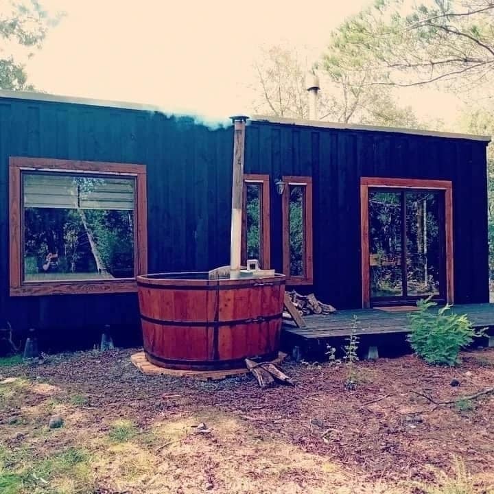 A wooden cabin features large windows framed in rich wood, allowing natural light to illuminate the interior. A traditional hot tub sits nearby, with steam rising gently into the fresh air. The surrounding area includes native trees and a natural landscape, enhancing the cabin's serene setting.