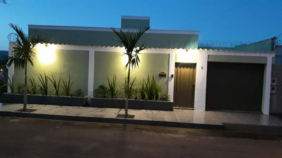 The exterior of the house features a modern façade with a light green color, accentuated by soft outdoor lighting. A well-maintained garden with palm trees and low shrubs borders the front, while a garage with a brown door is visible to one side.