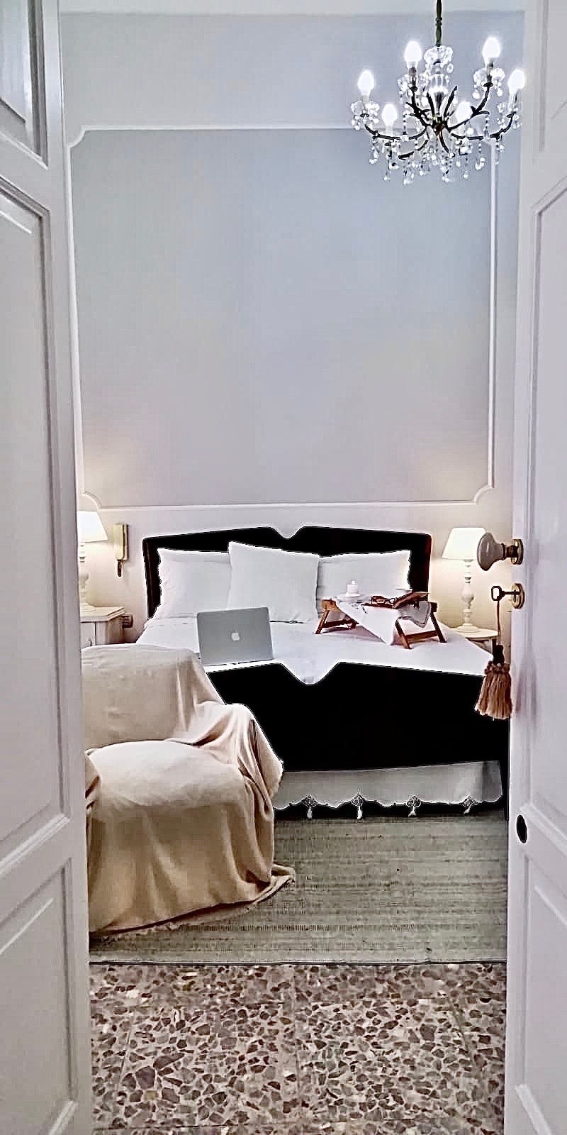 A well-appointed bedroom is visible through an open door, featuring a spacious bed with light linens and accent pillows. Soft lighting is provided by bedside lamps, and a small chandelier hangs from the ceiling. A cozy chair and decorative throw are placed beside the bed.