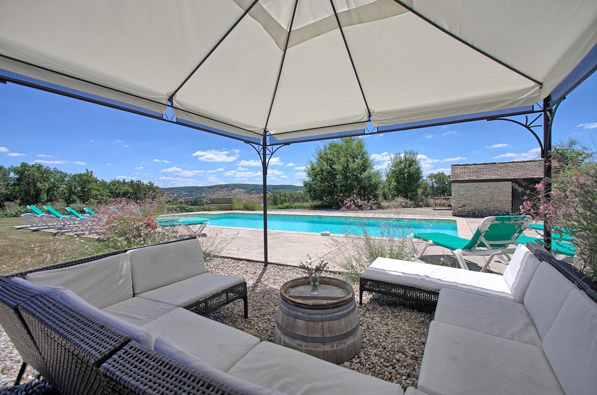 A shaded seating area is framed by a white canopy, featuring a circular table and a barrel at its center. In the background, a private swimming pool glimmers under the sun, surrounded by green loungers and open countryside views.