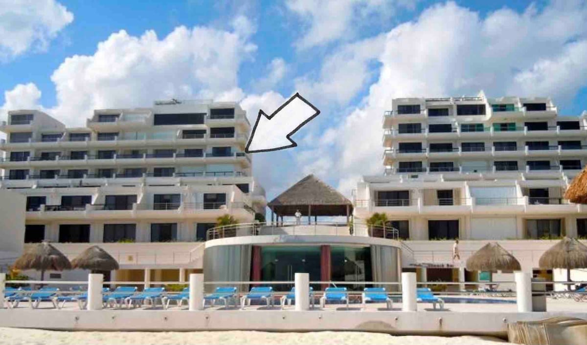A view of the beachfront complex features multiple levels with balconies overlooking the ocean. A thatched-roof structure is visible in the foreground, providing shade. Lounge chairs are arranged by the pool area, reflecting a relaxed atmosphere by the sea.