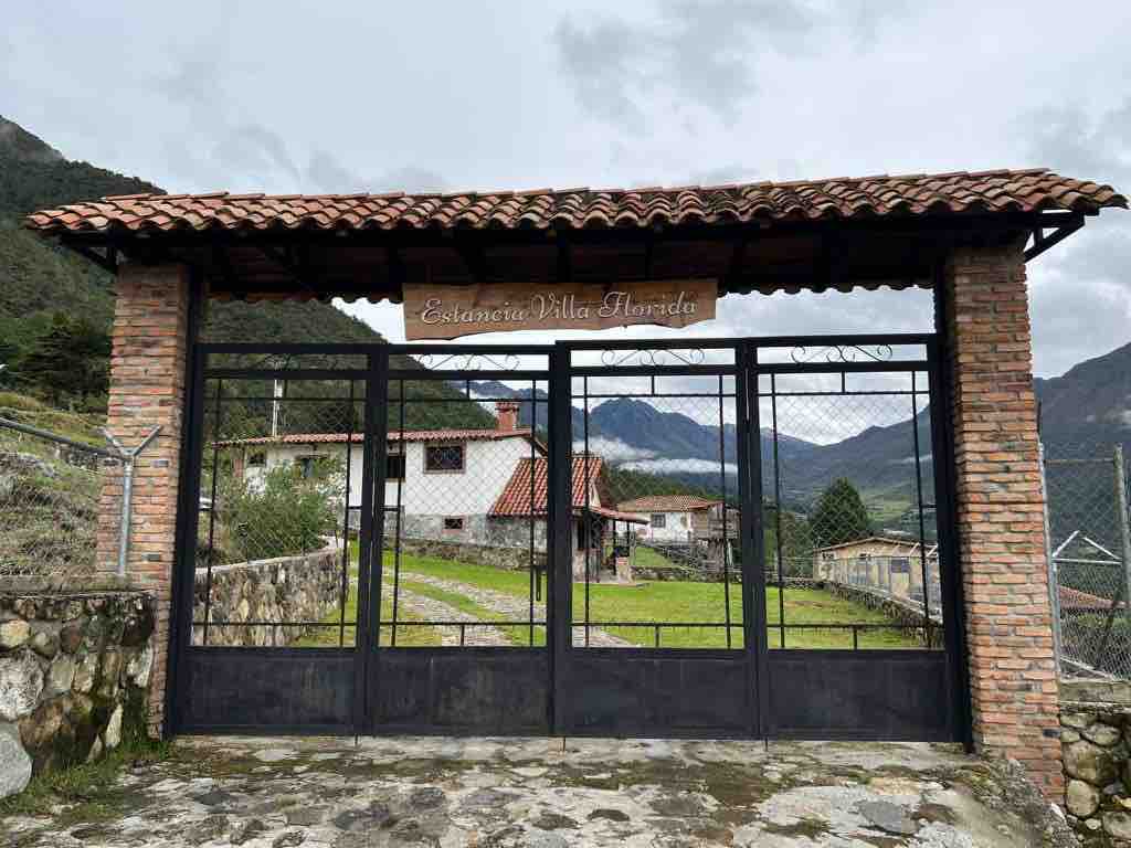 An inviting entrance gate of Estancia Villa Morada is framed by stone walls and topped with a traditional tile roof. Rolling green hills and mountains are visible in the background, providing a serene setting. A rustic pathway leads toward the inviting cabins beyond the gate.