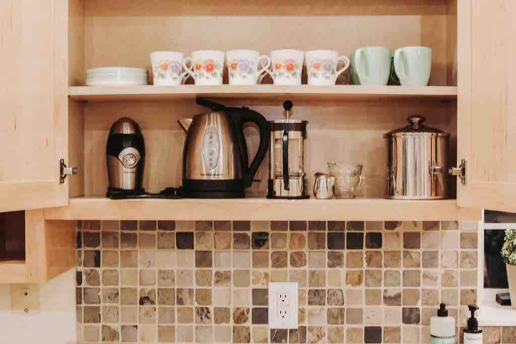 A kitchen shelf is organized with a collection of dinnerware, including floral-patterned cups and mint green mugs. An electric kettle, a French press, and a stainless-steel canister are positioned alongside a glass creamer. A textured stone backsplash complements the aesthetics.