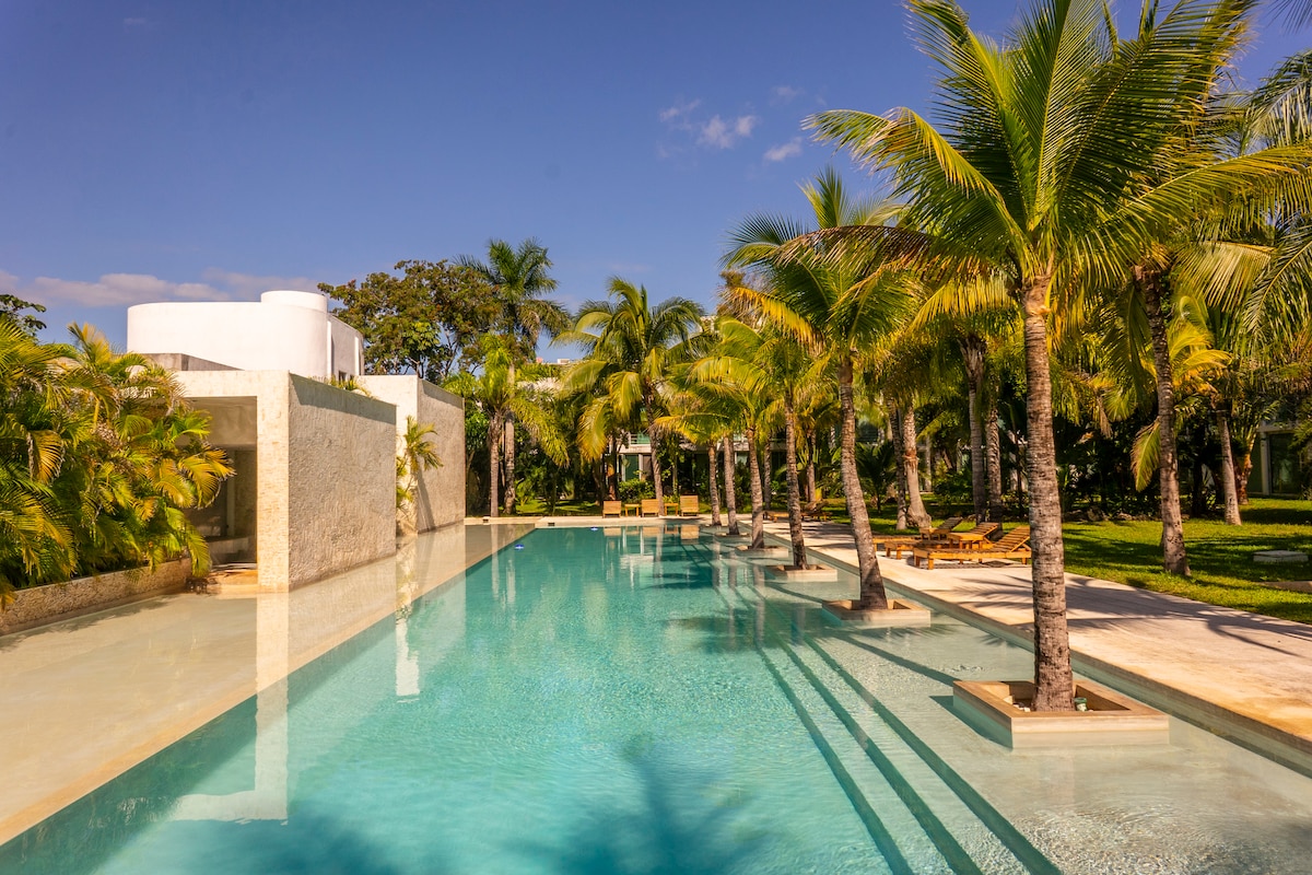 A large swimming pool is bordered by palm trees and lush tropical plants. Sun loungers are placed along the poolside, creating a serene outdoor atmosphere. The clear water reflects the blue sky and surrounding foliage.
