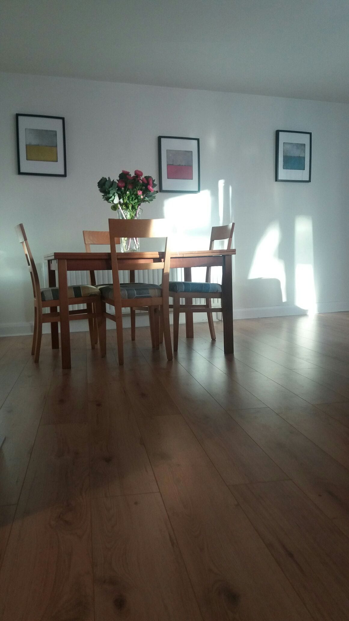 A dining area is presented with a wooden table and four matching chairs. A vase of roses sits in the center. Wall art consists of three framed pieces showcasing simple color patterns. Natural light from nearby windows creates soft shadows across the polished wooden floor.