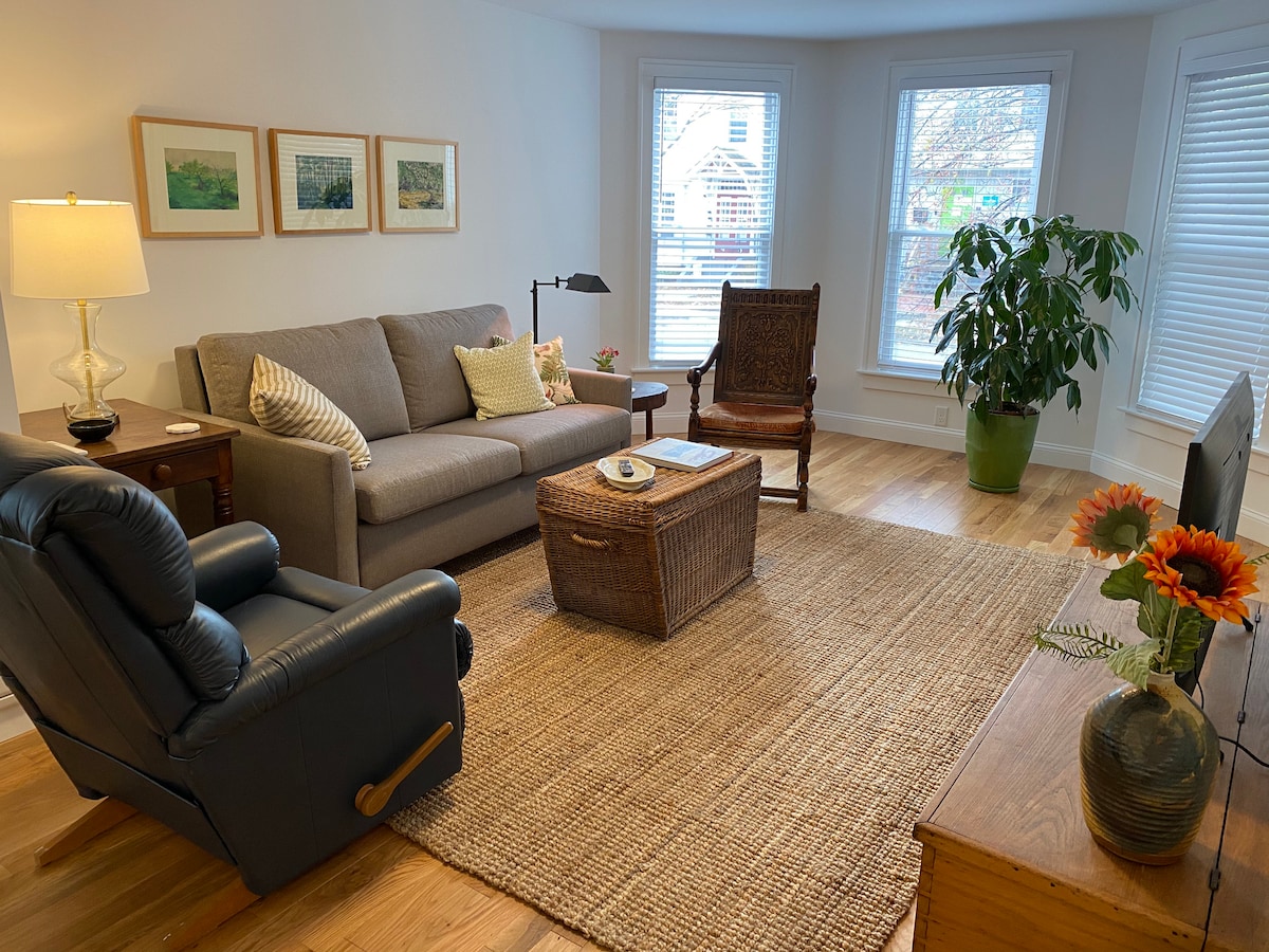 An open living area features a gray sofa with accent pillows, complemented by a wicker coffee table. A armchair is positioned nearby, with a standing lamp providing light. Large windows allow natural light to fill the space, alongside a green potted plant.